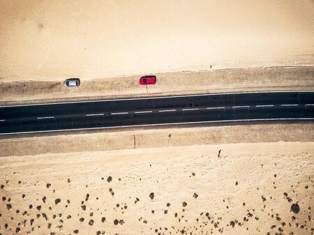 Aerial view of black straight asphalt road with sand and desert on both sides around - two car parked on the side - concept of travel and wanderlust for exotic destinationsの写真素材