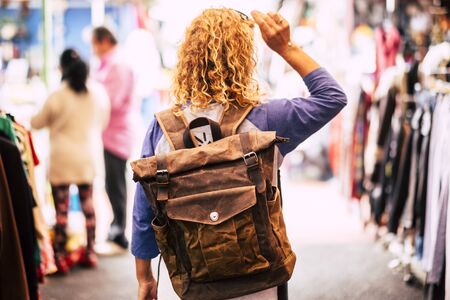 Blonde curly hair woman backpacker traveler viewed from rear at used market enjoying the shopping and alternative vacation - leather backpack view for travel conceptの写真素材