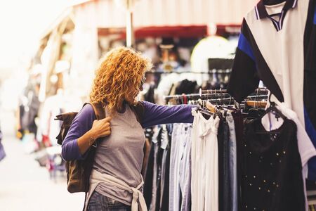 Cheerful traveler young blonde curly woman looking and choosing clothes at the used market during alternative vacation - shopping lifestyle concept for nice peopleの写真素材