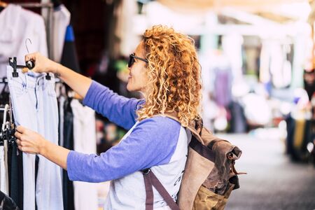 Cheerful traveler young blonde curly woman looking and choosing clothes at the used market during alternative vacation - shopping lifestyle concept for nice peopleの写真素材