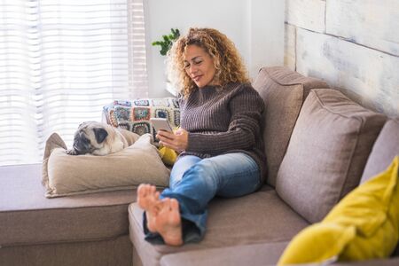 Happy adult beautiful curly woman at home typing and reading from celllar phone with her best friend dog pug relaxing at her side -  concept of lfiestyle for relaxed hapy independent peopleの写真素材