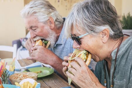 People eating hamburger junk food - Couple of senior man and woman with fast food lunch time - close up of elderly no healthy lifestyle - tasty sandwich full of everythingの写真素材