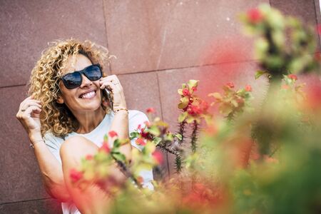 Cheerful adult young people woman talk at the phone sitting outdoor with flowers and red wall in background -female. and technology in leisure activity - independence and joyfulの写真素材