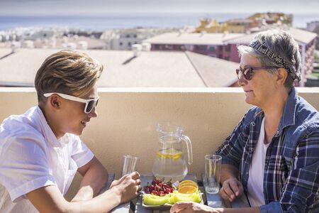 happy leisure activity on the terrace rooftop having breakfast with smiles and happiness for grandmother and teenager family caucasian people. ocean and buildings view, outdoor coule togetherの写真素材
