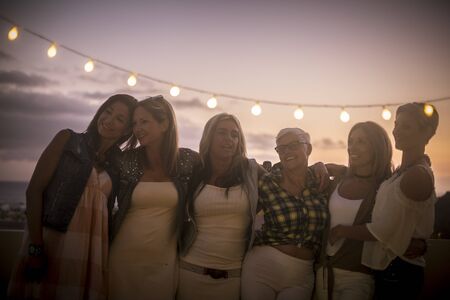 Romantic friendship concept with group of mixed generations and ages people caucasian women hug and enjoy the nightlife - sunset sky background and happy free young females togetherの写真素材