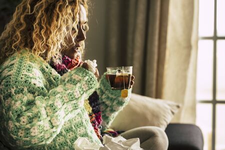 Adult beautiful caucasian woman at home sick with ill and cold drinking  hot tea - fever and seasonal influenza illness concept for unhealthy people sitting and taking medicineの写真素材