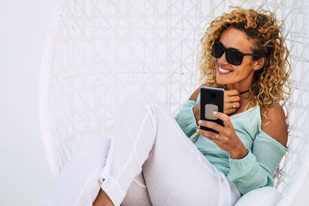 Cheerful beautiful curly young adult woman use mobile phone outdoor sit down on a white garden elegant chair -smile and enjoy the outdoor technology leisure activityの写真素材