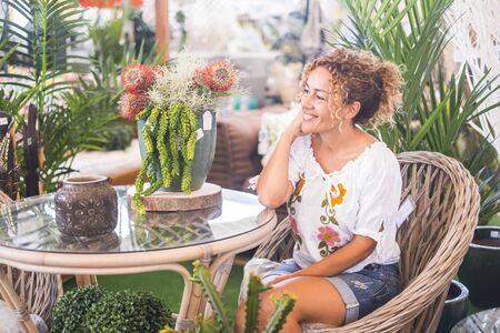 Young woman with blond curly hair sitting on a wicker chair with many plants admires the vegetation. Relaxation and leisure concept for people enjoying the nature and the environmentの写真素材