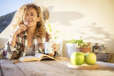 Cheerful relaxed beautiful curly blonde caucasianwoman enjoy the sunny day at home on the terrace reading a book and drinking tea or coffee from a mug - hipster style for pretty femaleの写真素材