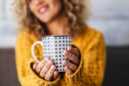 Close up of design trendy mug with tea or coffee or heahty drink holded by adult caucasian woman hands -  cheerful blonde female people in background defocusedの写真素材
