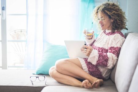 Pretty caucasian adult young woman at home drinking detox water and use a tablet device sitting on the sofa - females and technology at home with healthy lifestyle - beautiful curly hair lady relaxingの写真素材