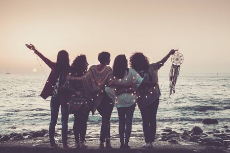 Love friendship and celebration time concept image with group of women friends viewed from back enjoying the beautiful coloured sunset in front of the ocean - feeling with nature and joyful for worldの写真素材