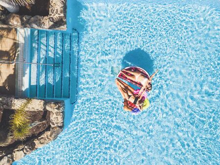 People having fun in summer at the pool with trendy coloured inflatable mattress lilos - couple of beautiful girls lay down and enjoy the sunny day together - aerial view and hotel resort concept vacationの写真素材