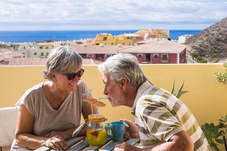 Retired people lifestyle with cheerful senior couple having fun together at home on the terrace drinking from the same glass a juice fruit - city and ocean view for vacation conceptの写真素材