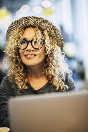 Beautiful adult cheerful happy woman work with laptop computer and bokeh coloured background - concept of technology and nice people with smart modern job - eyeglasses and blonde curly hairの写真素材