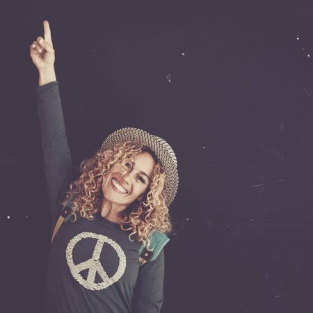 Cheerful beautiful caucasian adult woman with blonde curly hair and trendy hat sign with finger and smile at the camera for sale or offer message - black wall in background and peace on the shirtの写真素材
