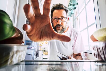 Adult caucasian man take healthy food from an open fridge - closeup inside view - weight loss concept -  home activity in the kitchen - quarantine coronavirus people concept stay homeの写真素材