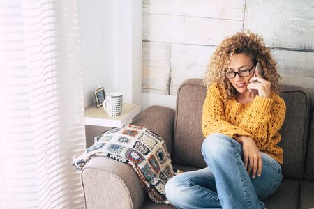 Beautiful adult people having phone call at home -curly caucasian blonde woman sitting on the sofa with cellular -  Middle age lady having nice time in relaxの写真素材