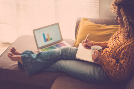 Beautiful caucasian adult woman working at home on the sofa with laptop and paper - concept of home...