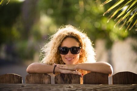 Portrait of beautiful caucasian young blonde curly woman cheerful looking at the camera with sun lights in background and defocused green natural outdoor background - happy middle age lady conceptの写真素材