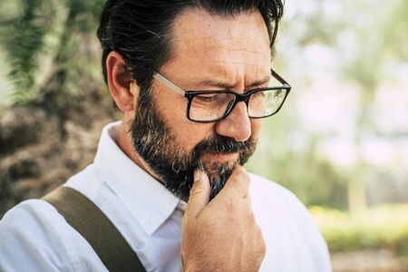 Worried adult caucasian 50 years old man touch the beard and think alone - eyeglasses and people outdoor with green park defocused backgroundの写真素材