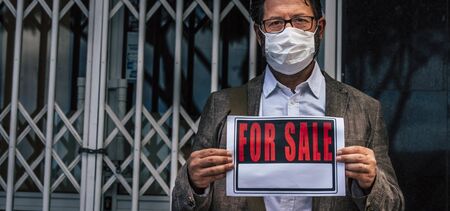 Concept of coronavirus covid-19 recession economy crisis with sad business man and for sale sign outside his store closed for lockdown phase  - people with mask protection healthの写真素材
