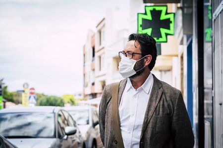 Adult mature caucasian man walk outdoor in the city using healthy protection mask for virus contagion and pharmacy in background - coronavirus new lifestyle conceptの写真素材
