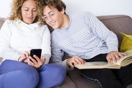Mother and son family enjoy time together sitting on the couch - young handsome teenager student with a book looking the mother modern phone - home lifestyleの写真素材