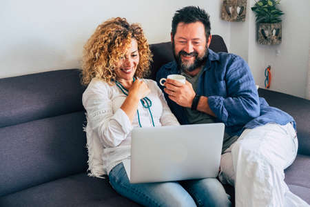 Happy adult aged couple caucasian people at home with personal laptop computer and internet connection - together in indoor leisure activity with technology sitting on the sofa and smilingの写真素材