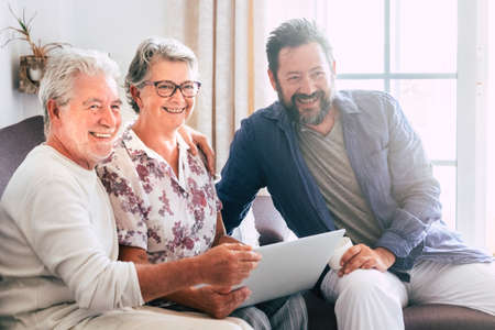 Cheerful happy mixed generation family at home with senior fathers and adult son smile and use a laptop computer sitting on the sofa - bright window backgroundの写真素材