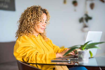 Adult pretty woman at home work with laptop computer on the table- concept of modern smart woking and job alternative office - blonde curly hair and beutiful people in technology internet connected activityの写真素材