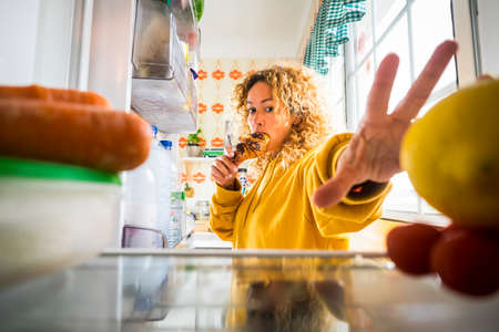 Hungry beautiful adult young woman eat sweet cake croissant and open the fridge to look for other food - diet and food concept - view from inside the fridgeの写真素材