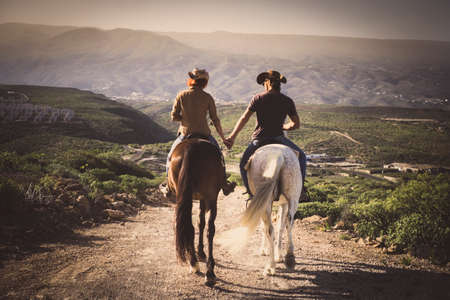 Romantic couple wide horses together holding hands with love and romance in outdoor leisure lifestyle alternative activity - viewed from back young people with aninmalsの写真素材