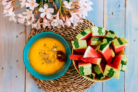 Above colored food view with yellow vegetables soup and red fresh watermelon fruit on a blue yellow table with flowers decorationの写真素材