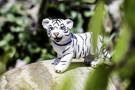 Little White Bengak Tiger Figurine standing on a grey rock with beautiful green foilage in the backgroundの写真素材