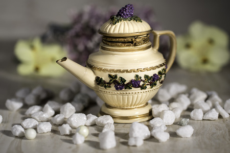 Miniature Beige Teapot with lid and grapevine decorations against a flower background surrounded by small white stones.の写真素材