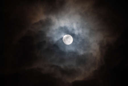 Half-hidden full moon in a cloudy night sky.の写真素材