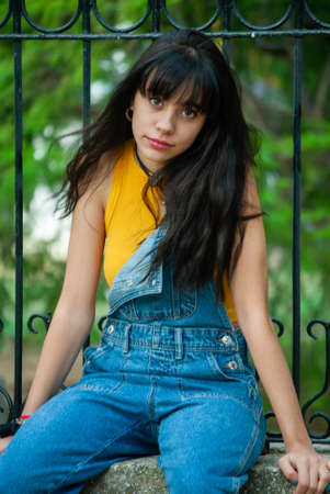 Young girl dressed in yellow and wearing a denim jumpsuit sitting on a stone wall in a park with autumn green and yellow colors.の写真素材