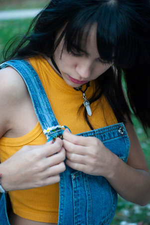 Young girl dressed in yellow putting some daisies on her denim overalls in a park with autumn green and yellow colors.の写真素材