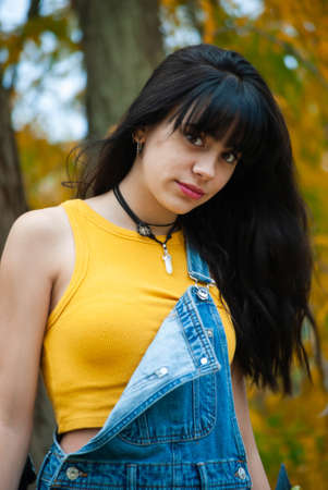Young girl dressed in yellow and wearing a denim jumpsuit posing in a park with autumn green and yellow colorsの写真素材