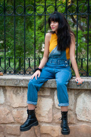 Young girl dressed in yellow and wearing a denim jumpsuit sitting on a stone wall in a park with autumn green and yellow colorsの写真素材