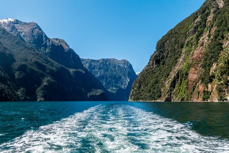 Views on the waters of Milford Sound with the rare sight of the fjord in sunshine. Snow capped mountains in the background fall off steeply into the sea.の写真素材