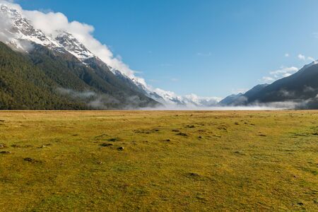 In the early morning fog covers the valley ground while the sun illuminates the snow covered mountains at both sides. A vast, flat grass plain covers the flat valley floor.の写真素材