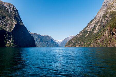 Views on the waters of Milford Sound with the rare sight of the fjord in sunshine. Snow capped mountains in the background fall off steeply into the sea.の写真素材
