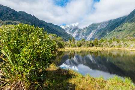 Snow covered mountains mirror in a little lake on New Zealand's West coast. Dark clouds nearly cover the peaks while temperate rainforests cover the slopes of the mountains.の写真素材
