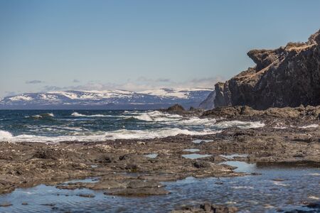 Snow capped hills in the distance at sunset against a clear blue sky. White rapids and rocks in the sea. Large craggy cliffside. Gros Morne National Park, Newfoundland, Canada.の写真素材