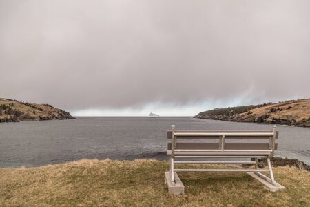 Large iceberg on the sea under storm clouds in Torbay, Newfoundland, Canada. A park bench sits in the forefront on yellow and green grass.の写真素材