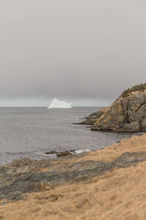 Large iceberg on the sea under storm clouds in Torbay, Newfoundland, Canada. Yellow grass and rocks in the forefront.の写真素材