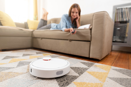 Robotic vacuum cleaner cleaning the room while woman relaxing on sofa. Woman controlling vacuum with remote control.の写真素材