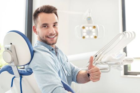 Handsome man showing thumbs up and smiling sitting at the dental chair. Professional dental clinic, healthy teeth concept.の写真素材